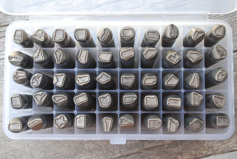 May include: A clear plastic storage case filled with 48 metal stamps, each featuring the outline of a different US state. The stamps are arranged in rows and columns, and the case has a clear lid.