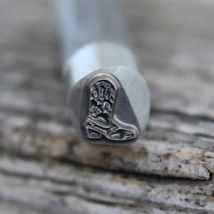 Puede incluir: Un sello de metal con un diseño de una bota vaquera. La bota está detallada con costuras y una punta puntiaguda.