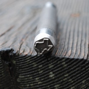 May include: A close-up of a metal stamp with a design of the state of Texas. The stamp is silver and has a textured surface.