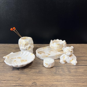May include: A set of five white concrete home decor items with gold accents. The set includes a small vase, a tray, a small bowl, a candle holder, and a small bear figurine.