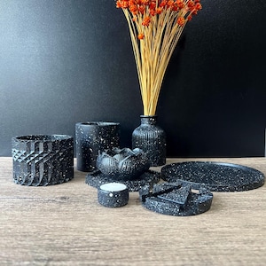 Puede incluir: Una colección de artículos de decoración para el hogar negros moteados, que incluyen un jarrón con flores secas naranjas, una maceta geométrica, un portavelas en forma de loto, una bandeja redonda y un pequeño portavelas. Los artículos están dispuestos sobre una superficie de madera.