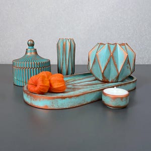 Puede incluir: Colección de objetos decorativos de color turquesa y cobre. Incluye un tarro con tapa, dos jarrones geométricos, una bandeja con dos flores naranjas, un cuenco geométrico y una vela pequeña. Los objetos tienen un aspecto envejecido.