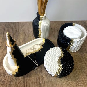 Puede incluir: Colección de accesorios decorativos para el hogar en negro, blanco y dorado. Incluye un portaanillos, una bandeja, un jarrón con hierba seca y un recipiente pequeño. Los artículos tienen un diseño moderno con formas geométricas y detalles dorados.