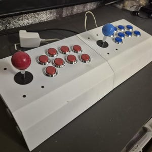 Op de afbeelding: Een witte arcadecontroller met een rode joystick en rode knoppen aan de ene kant, en een blauwe joystick en blauwe knoppen aan de andere kant. De controller heeft een rechthoekige vorm.