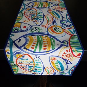 Könnte beinhalten: Ein weißer Tischläufer mit blauem Rand zeigt ein farbenfrohes Muster aus stilisierten Fischen in Blautönen, Grün, Gelb, Orange und Rot. Die Fische sind in einem skurrilen Stil mit abstrakten Details gemalt.