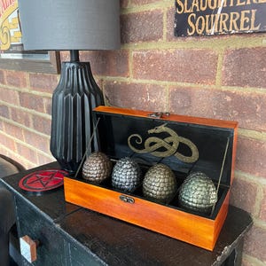 May include: A rectangular wooden box with a hinged lid, containing four textured, egg-shaped objects in bronze, silver, gold, and green finishes. The interior lid features a gold-coloured dragon emblem, and the box is secured with a metal latch.