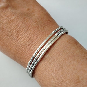 May include: A set of three silver bracelets. Two bracelets have a textured, ridged design, while the third features small, round beads. The bracelets are worn on a wrist, showcasing their simple yet elegant design. The bracelets are made of a shiny, metallic material.