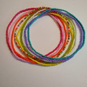 Op de afbeelding: Een verzameling kleurrijke kralenkettingen in verschillende tinten, waaronder roze, oranje, geel, groen, paars en blauw. De kettingen zijn in een cirkelvormig patroon gerangschikt, waarbij de levendige kleuren en het ingewikkelde kralenwerk worden getoond. Sommige kettingen hebben goudkleurige kralen.