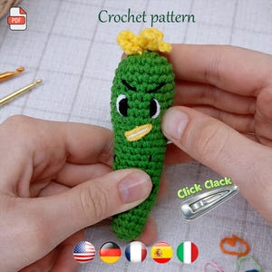 May include: A handmade green crochet pickle with black eyes, a yellow beak, and a yellow flower on top. The text "Crochet pattern" is above the pickle. A PDF icon is in the upper left corner.