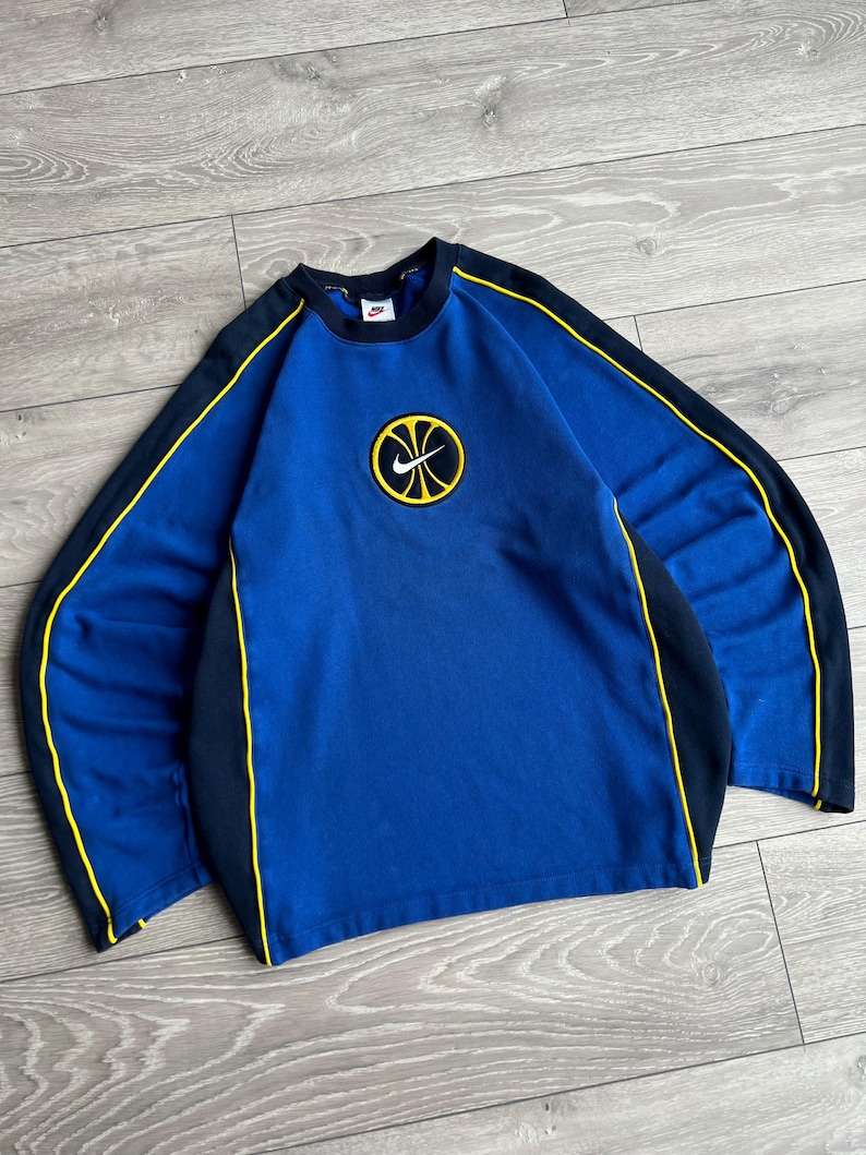 May include: A blue sweatshirt with black and yellow accents. The sweatshirt features a basketball logo on the chest and black stripes with yellow trim along the sleeves. The Nike logo is visible on the upper chest.