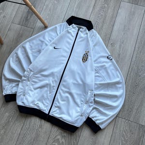 May include: White zip-up jacket with black trim and a team logo on the chest. Features the Nike swoosh logo on the chest and sleeve. The jacket has two front pockets and a black collar.