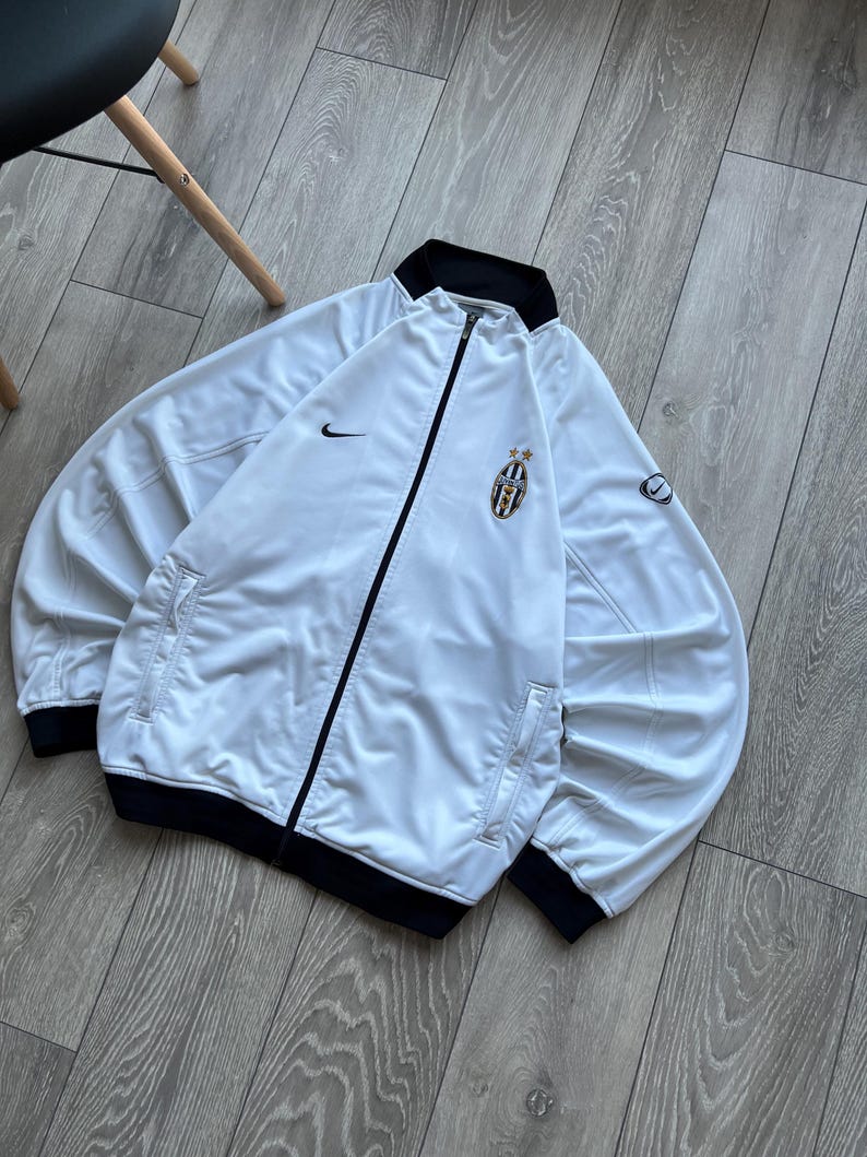 May include: White zip-up jacket with black trim and a team logo on the chest. Features the Nike swoosh logo on the chest and sleeve. The jacket has two front pockets and a black collar.