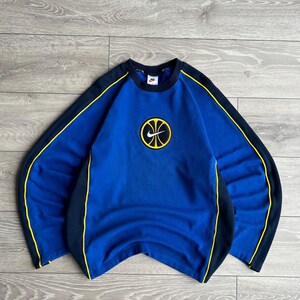 May include: Blue Nike sweatshirt with a basketball logo. The sweatshirt has a crew neck and long sleeves. It features black and yellow accents along the sleeves and sides. The Nike logo is visible on the chest.