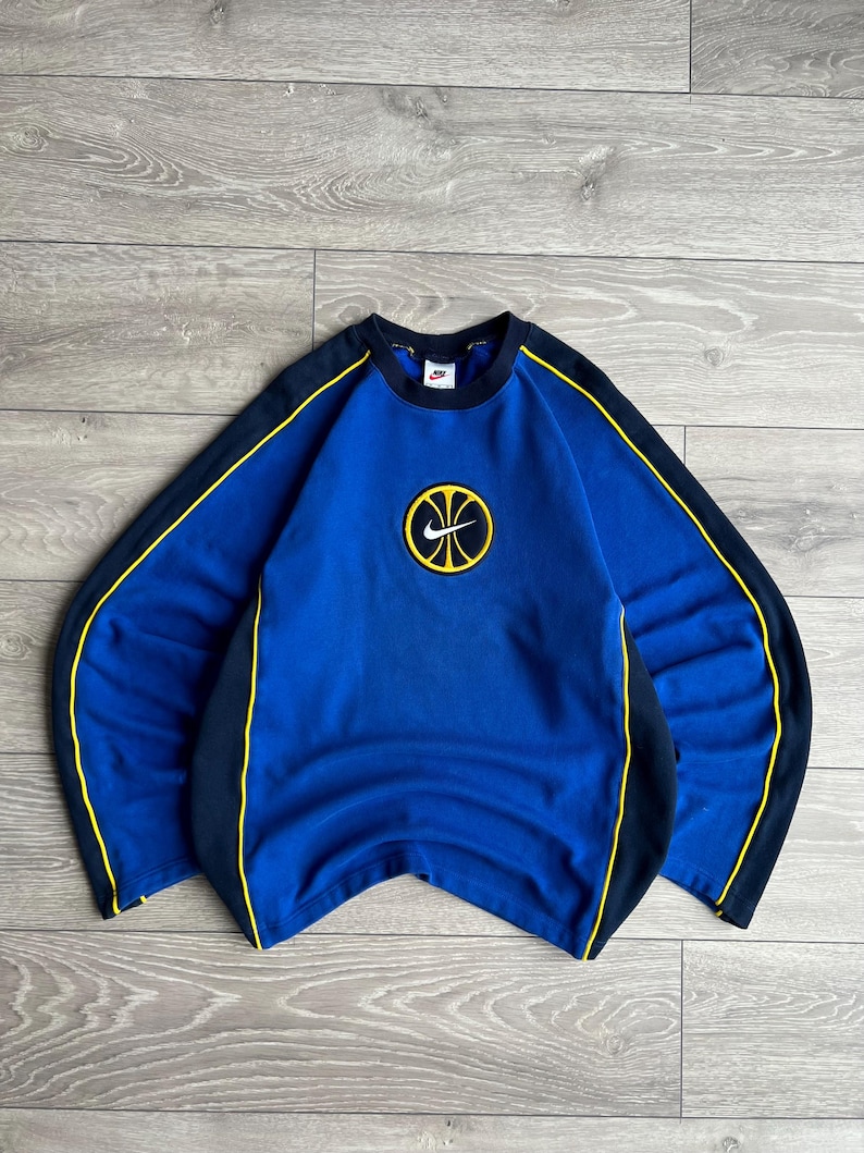 May include: Blue Nike sweatshirt with a basketball logo. The sweatshirt has a crew neck and long sleeves. It features black and yellow accents along the sleeves and sides. The Nike logo is visible on the chest.