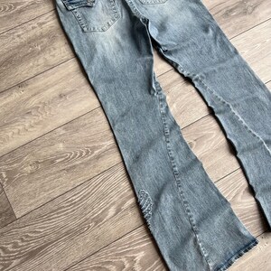 May include: Light blue denim jeans featuring a flared leg silhouette, two back pockets with button-down flaps, and a decorative beaded wing-shaped embellishment on the lower left leg. The jeans have a distressed wash and a frayed waistband edge.