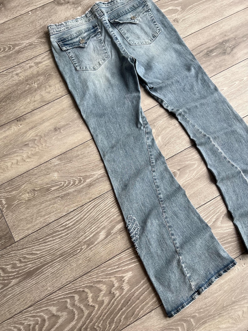 May include: Light blue denim jeans featuring a flared leg silhouette, two back pockets with button-down flaps, and a decorative beaded wing-shaped embellishment on the lower left leg. The jeans have a distressed wash and a frayed waistband edge.