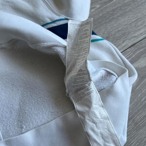 Puede incluir: Sudadera con capucha blanca con detalle de rayas azules y turquesas. El interior es de tejido blanco suave. Una etiqueta blanca con texto est&aacute; adherida a la sudadera.