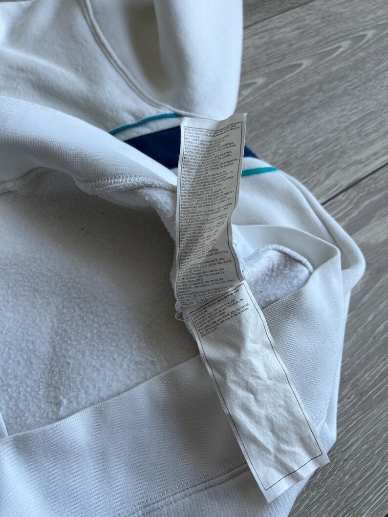 Puede incluir: Sudadera con capucha blanca con detalle de rayas azules y turquesas. El interior es de tejido blanco suave. Una etiqueta blanca con texto est&aacute; adherida a la sudadera.