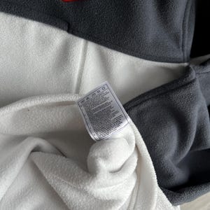 May include: A grey and white fleece pullover with a small red Nike logo. The garment has a tag with care instructions. The fleece is soft and appears to be new.