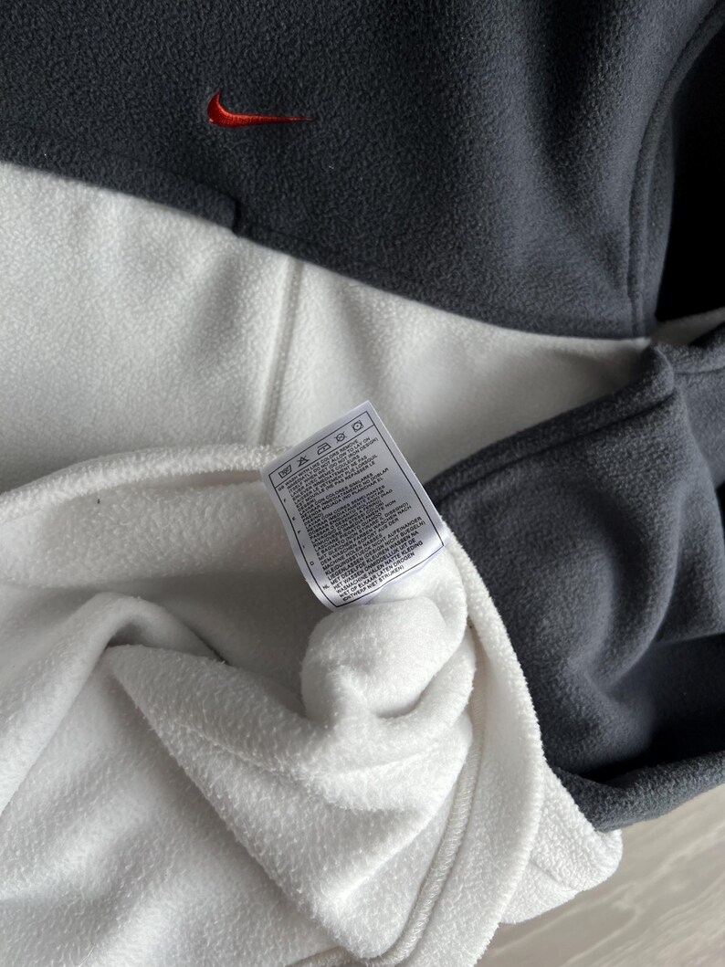 May include: A grey and white fleece pullover with a small red Nike logo. The garment has a tag with care instructions. The fleece is soft and appears to be new.