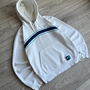 Puede incluir: Sudadera con capucha blanca con bolsillo delantero y una franja azul y turquesa en el pecho. El logo de Nike es visible en la parte superior izquierda. Un peque&ntilde;o parche est&aacute; en la parte inferior derecha.