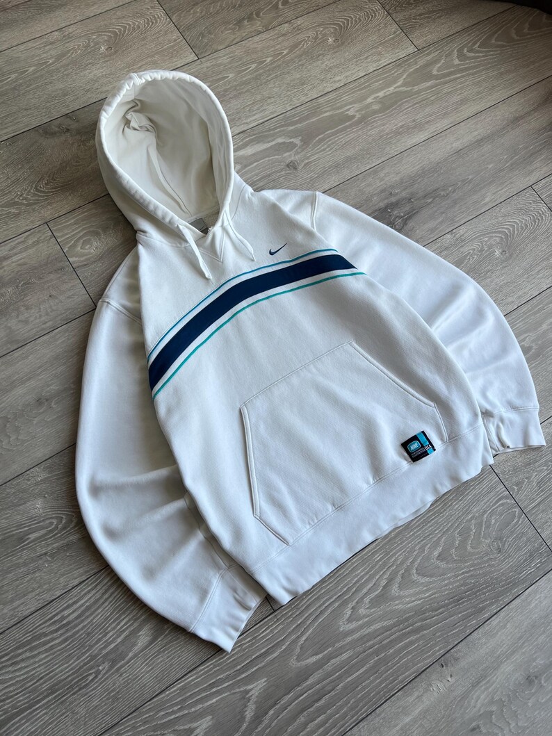 Puede incluir: Sudadera con capucha blanca con bolsillo delantero y una franja azul y turquesa en el pecho. El logo de Nike es visible en la parte superior izquierda. Un peque&ntilde;o parche est&aacute; en la parte inferior derecha.