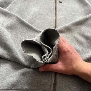 Puede incluir: Sudadera gris con cremallera y logotipo blanco de Nike en el pecho. La sudadera con capucha tiene una capucha con cord&oacute;n y pu&ntilde;os y dobladillo acanalados. La cremallera es plateada. Una mano sostiene la manga.