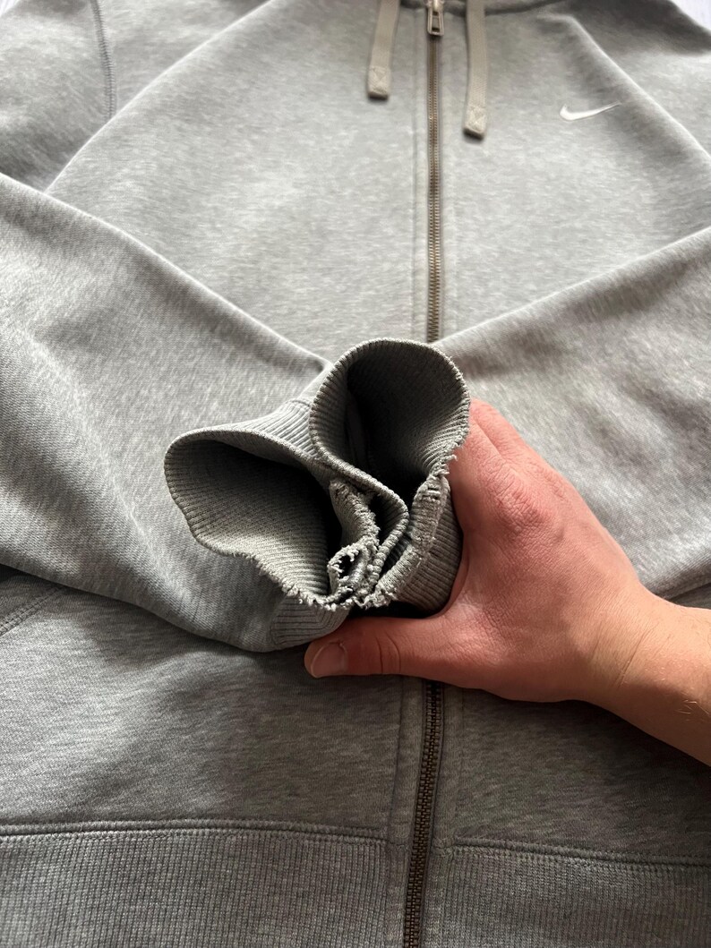 Puede incluir: Sudadera gris con cremallera y logotipo blanco de Nike en el pecho. La sudadera con capucha tiene una capucha con cord&oacute;n y pu&ntilde;os y dobladillo acanalados. La cremallera es plateada. Una mano sostiene la manga.