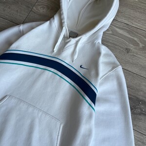 Puede incluir: Sudadera con capucha blanca con una franja azul y turquesa en el pecho. Cuenta con un bolsillo delantero y el logo de Nike swoosh. La sudadera tiene cordones y un ajuste relajado.