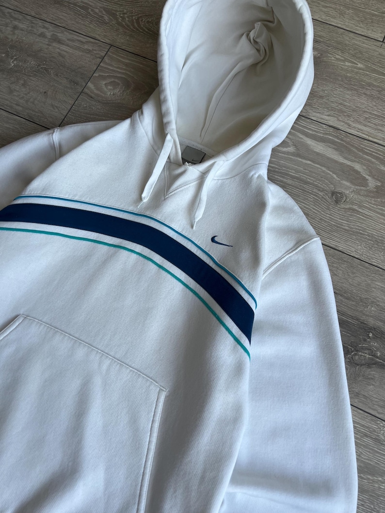 Puede incluir: Sudadera con capucha blanca con una franja azul y turquesa en el pecho. Cuenta con un bolsillo delantero y el logo de Nike swoosh. La sudadera tiene cordones y un ajuste relajado.