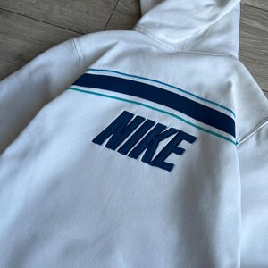 Puede incluir: Sudadera con capucha blanca con un dise&ntilde;o de rayas azul marino y turquesa en la espalda. La palabra "NIKE" est&aacute; bordada en azul marino en la parte trasera de la sudadera.