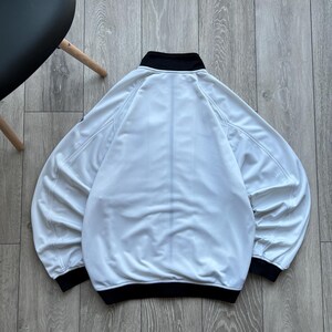 May include: A white jacket with a black collar and cuffs. The jacket has a full zip and long sleeves. The jacket is made of a soft, lightweight material. The jacket is shown from the back.