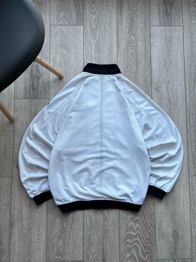 May include: A white jacket with a black collar and cuffs. The jacket has a full zip and long sleeves. The jacket is made of a soft, lightweight material. The jacket is shown from the back.