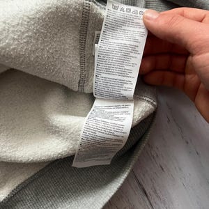 Puede incluir: Primer plano del interior de una sudadera gris con forro blanco y difuso. Se ve una etiqueta con instrucciones de cuidado. La prenda est&aacute; hecha de un material suave y texturizado, adecuado para uso casual.