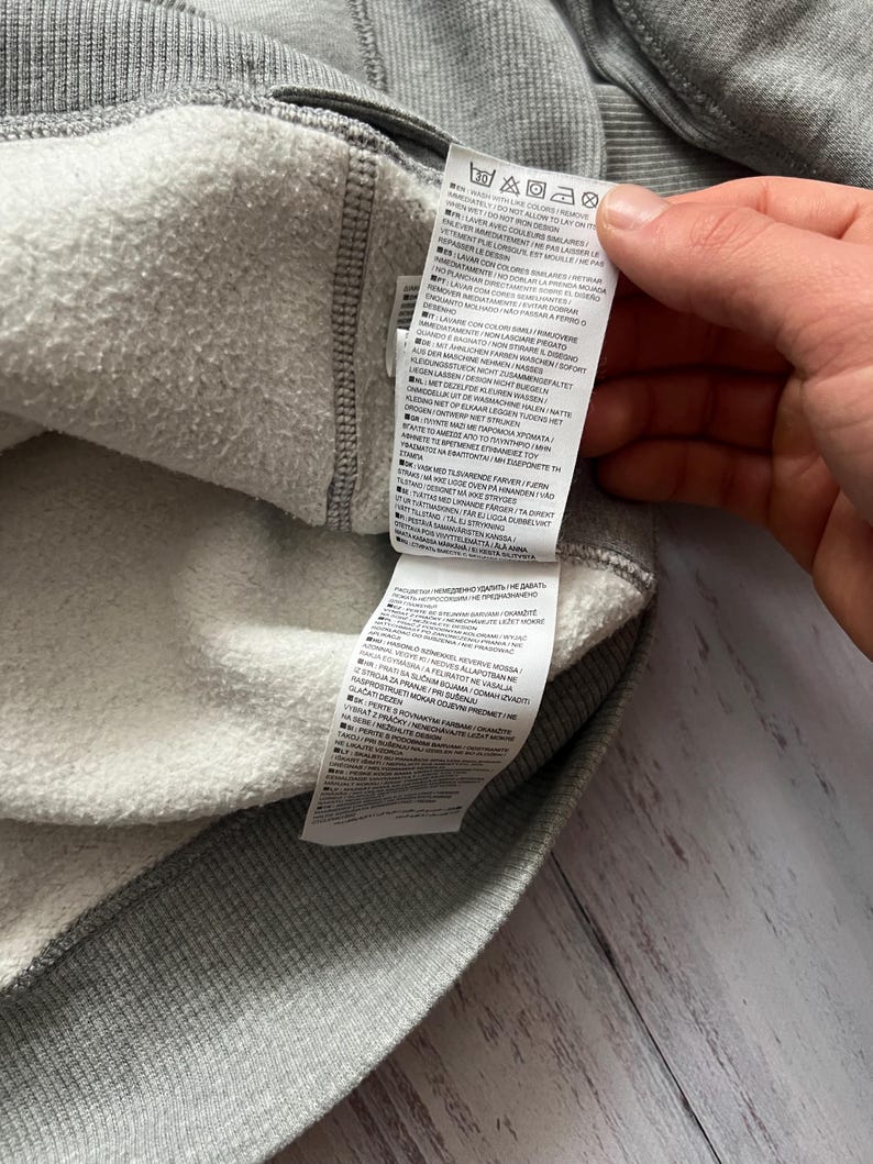 Puede incluir: Primer plano del interior de una sudadera gris con forro blanco y difuso. Se ve una etiqueta con instrucciones de cuidado. La prenda est&aacute; hecha de un material suave y texturizado, adecuado para uso casual.