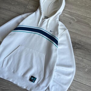 Puede incluir: Sudadera con capucha Nike blanca con una franja turquesa y azul marino en el pecho. Cuenta con una capucha con cordones, un bolsillo delantero y el logo de Nike. Una peque&ntilde;a etiqueta es visible en la parte inferior izquierda.