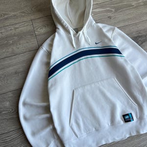 Puede incluir: Sudadera con capucha blanca con bolsillo delantero y una franja azul y turquesa en el pecho. El logotipo de Nike es visible en la parte superior izquierda. Una peque&ntilde;a etiqueta est&aacute; en la parte inferior derecha.