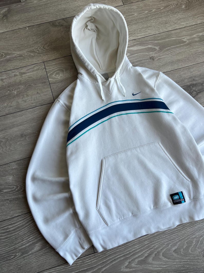 Puede incluir: Sudadera con capucha blanca con bolsillo delantero y una franja azul y turquesa en el pecho. El logotipo de Nike es visible en la parte superior izquierda. Una peque&ntilde;a etiqueta est&aacute; en la parte inferior derecha.