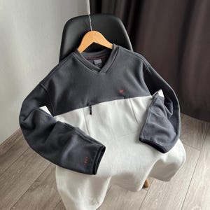 May include: A two-tone fleece pullover in charcoal grey and white, featuring a V-neck and a small red logo. The long-sleeved top has a small zip and a red logo on the sleeve.