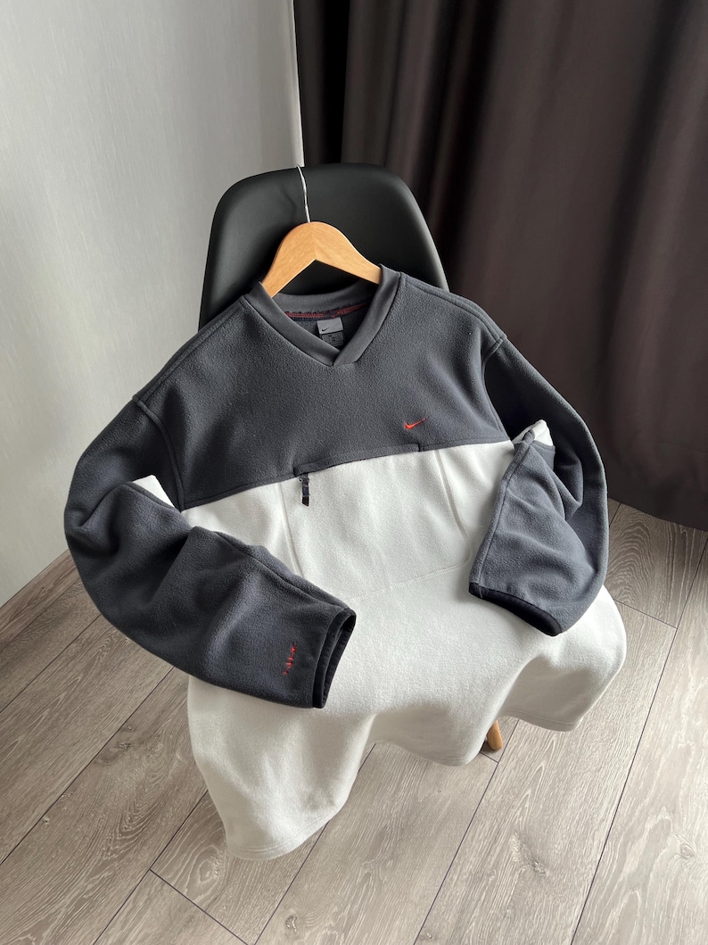 May include: A two-tone fleece pullover in charcoal grey and white, featuring a V-neck and a small red logo. The long-sleeved top has a small zip and a red logo on the sleeve.