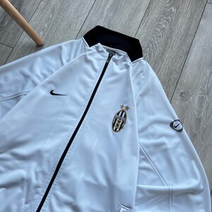 May include: White zip-up jacket with black trim and a black collar. Features the Nike swoosh logo and a team emblem with stars. The jacket is made of a smooth, lightweight material.