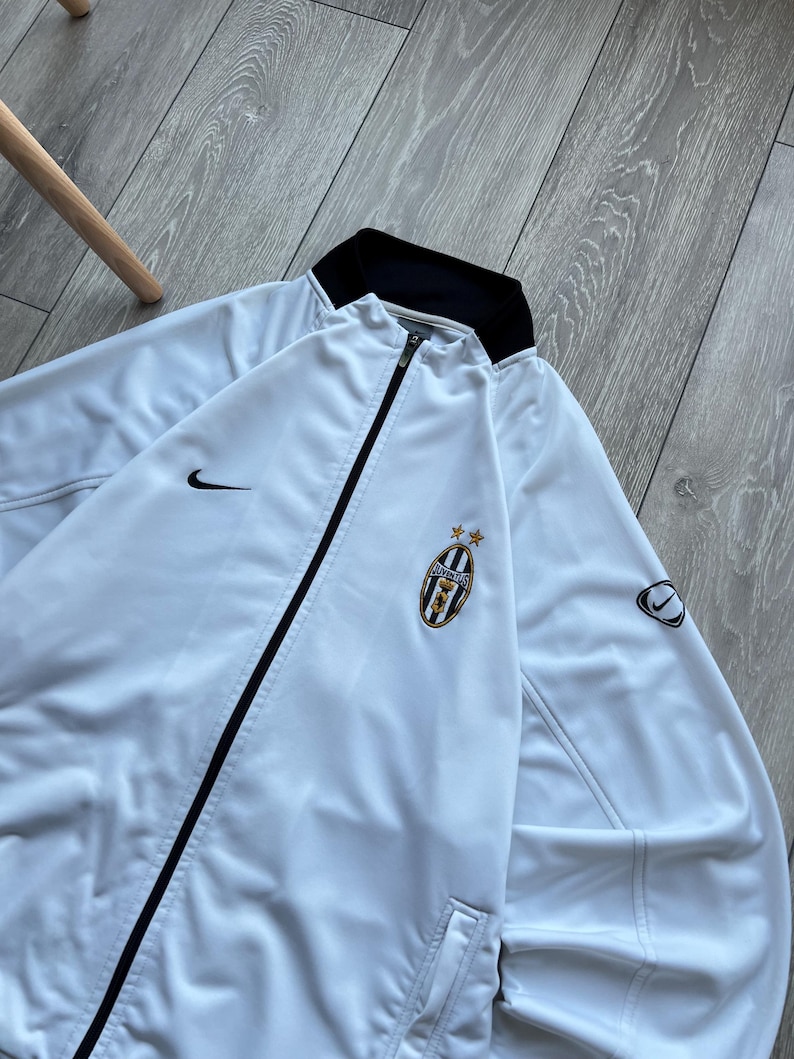 May include: White zip-up jacket with black trim and a black collar. Features the Nike swoosh logo and a team emblem with stars. The jacket is made of a smooth, lightweight material.