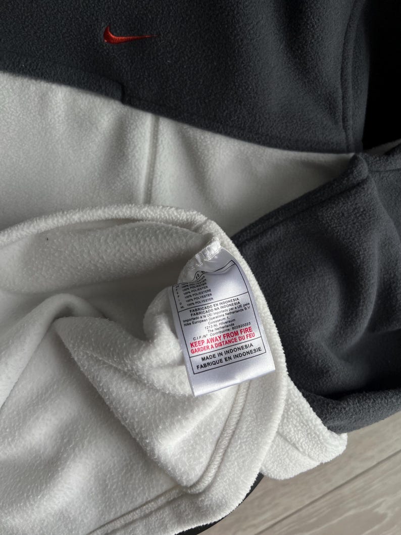 May include: A grey and white fleece pullover with a small red Nike logo. The garment has a tag that reads "KEEP AWAY FROM FIRE" and "MADE IN INDONESIA".