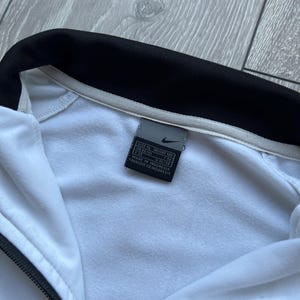 May include: White Nike track jacket with a black collar and zip. The jacket has a tag with the Nike logo and text. Made in Indonesia.