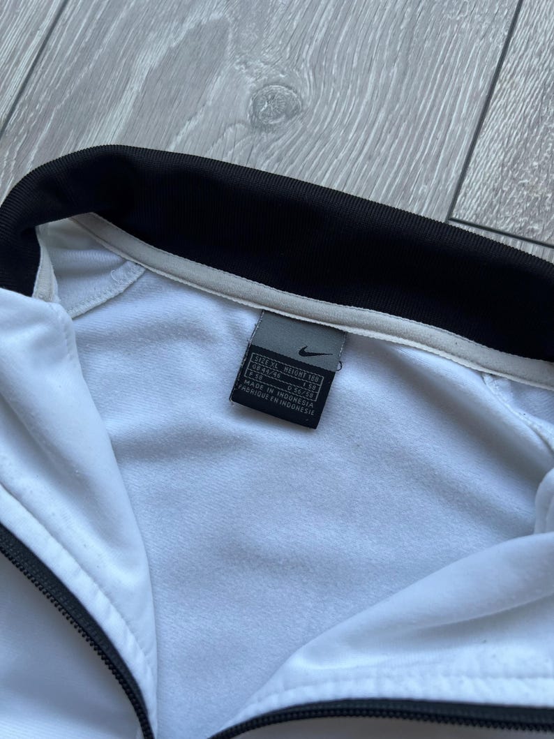 May include: White Nike track jacket with a black collar and zip. The jacket has a tag with the Nike logo and text. Made in Indonesia.