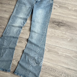 May include: Light blue denim jeans featuring a flared leg silhouette, a frayed waistband edge, and a decorative embroidered wing design in light blue thread on the lower left leg. The jeans include a front button closure and classic pockets.
