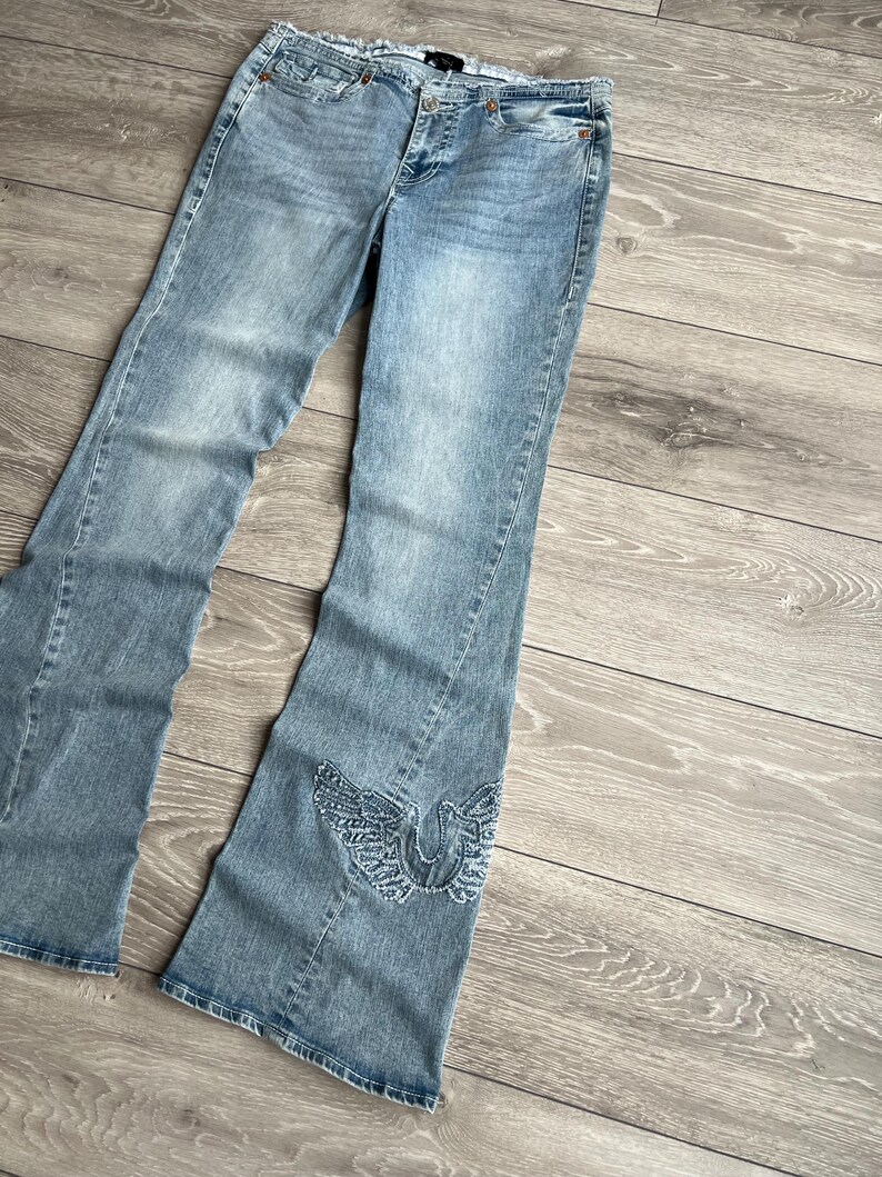 May include: Light blue denim jeans featuring a flared leg silhouette, a frayed waistband edge, and a decorative embroidered wing design in light blue thread on the lower left leg. The jeans include a front button closure and classic pockets.