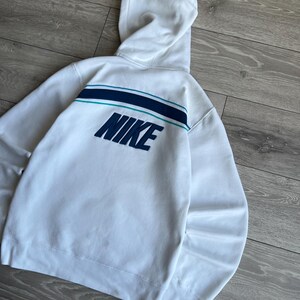 Puede incluir: Sudadera con capucha blanca con el logo de Nike en azul marino en la espalda. La sudadera tiene una capucha y un dise&ntilde;o de rayas horizontales en azul marino y turquesa. La prenda est&aacute; hecha de un material suave.