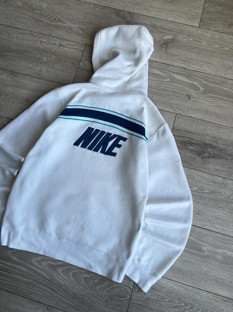Puede incluir: Sudadera con capucha blanca con el logo de Nike en azul marino en la espalda. La sudadera tiene una capucha y un dise&ntilde;o de rayas horizontales en azul marino y turquesa. La prenda est&aacute; hecha de un material suave.