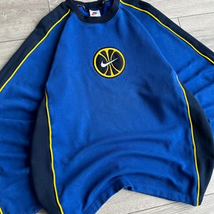 May include: A blue sweatshirt with a round Nike logo featuring a basketball design. The sweatshirt has black and yellow accents on the sleeves and sides. The Nike logo is centered on the chest.
